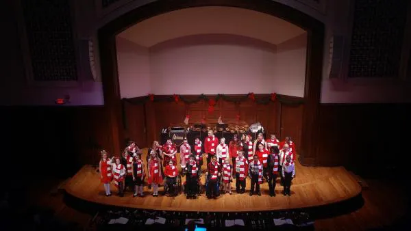 A wide shot photo of the holiday concert where a group of people are standing on stage singing while wearing red and white scarves.
