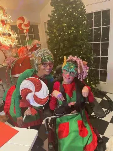 A woman and a kid in their wheelchair are dressed up in Christmas outfits and holding a huge peppermint lollipop.