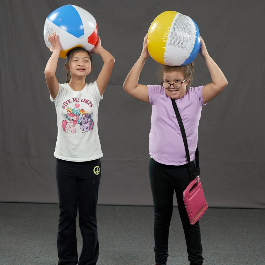 two young girls are standing next two each other smiling and holding inflatable beach balls above their heads.
