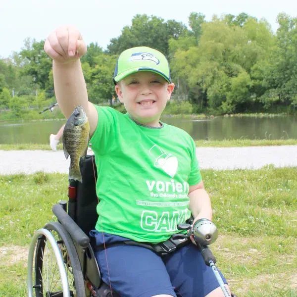 A young boy is holding a fish he caught while sitting n his wheelchair at camp.